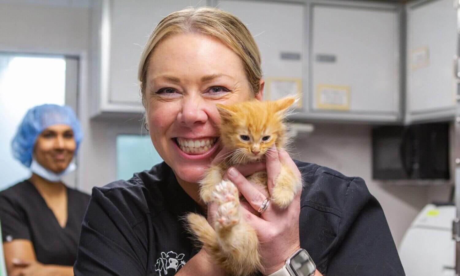 A woman in black scrubs smiles and holds an orange kitten, showcasing the joy of their work. A person in the background wears a hairnet and scrubs in a clinical setting, where 70,000 spay/neuter surgeries are performed annually.