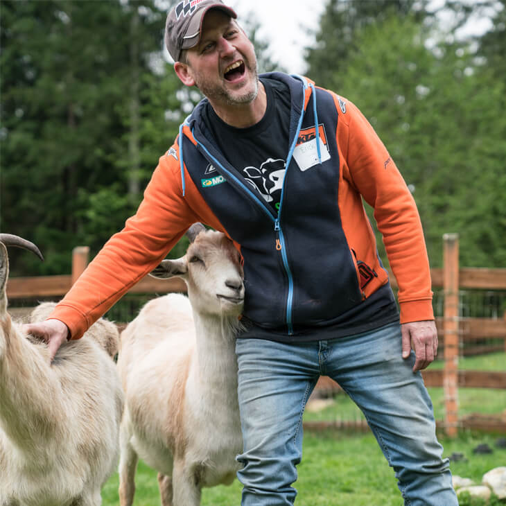 A man in an orange jacket stands in a grassy area with two goats, smiling and looking up, enjoying the peaceful atmosphere of an animal sanctuary tour.