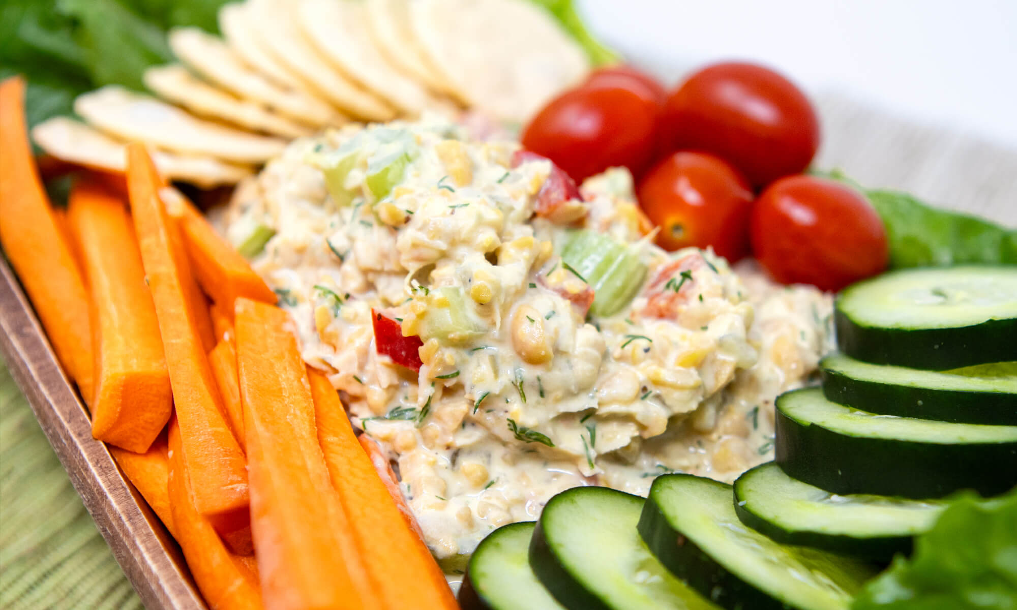 A platter with sliced carrots, cucumber, cherry tomatoes, round crackers, and a chickpea salad.