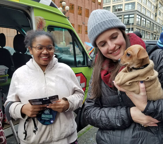 Two people stand near a green van, embodying inclusion and diversity. One holds a small dog in a brown sweater, while the other carries an informative pamphlet. Both are smiling warmly.