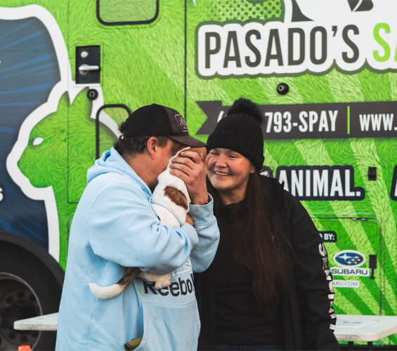 A person in a blue hoodie holds a small dog close, while another in a black jacket smiles at them.