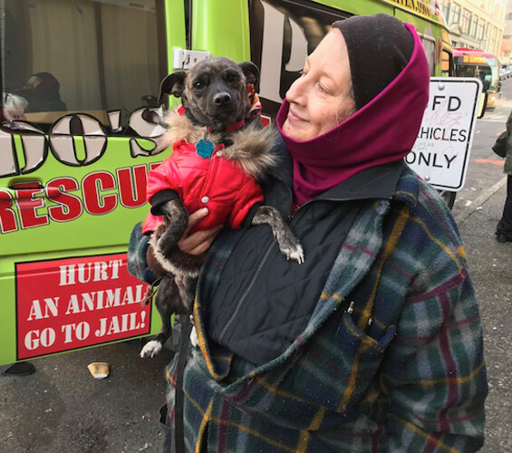 A person in a headscarf holds a small dog wearing a red jacket, standing proudly in front of an animal rescue vehicle.