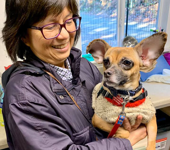 A woman wearing glasses and a dark jacket smiles while holding a small dog in a sweater indoors.