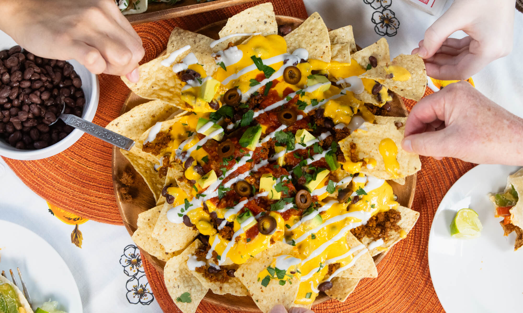 Three hands reach for loaded nachos topped with cheese, salsa, olives, and cilantro. A bowl of black beans and other dishes complete this budget-friendly vegan spread—perfect inspiration for your next vegan cooking adventure or free cooking class.