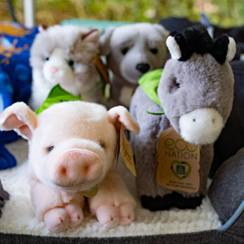 Four plush animal toys—a pig, cat, dog, and donkey—sit close together, each wearing a small green scarf with eco-friendly tags attached, making them a charming choice for charitable giving or as thoughtful ways to give back.