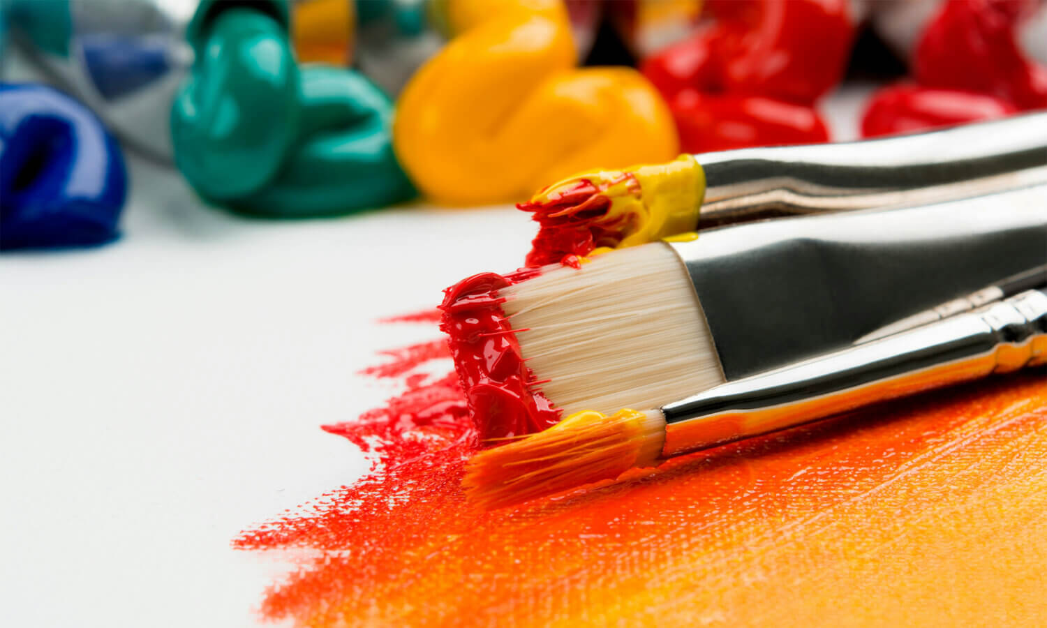Three paintbrushes with red and yellow paint rest on a canvas, blending colors, with tubes of green, yellow, and blue paint in the background.