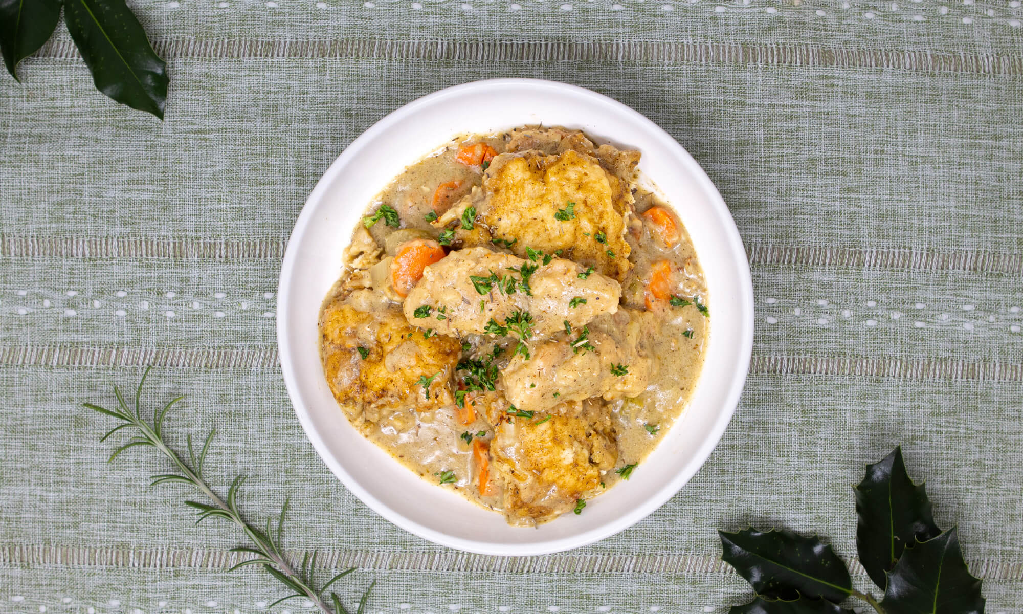 A white bowl filled with vegan chicken and vegetable stew topped with dumplings, garnished with chopped herbs, sits on a green patterned tablecloth.