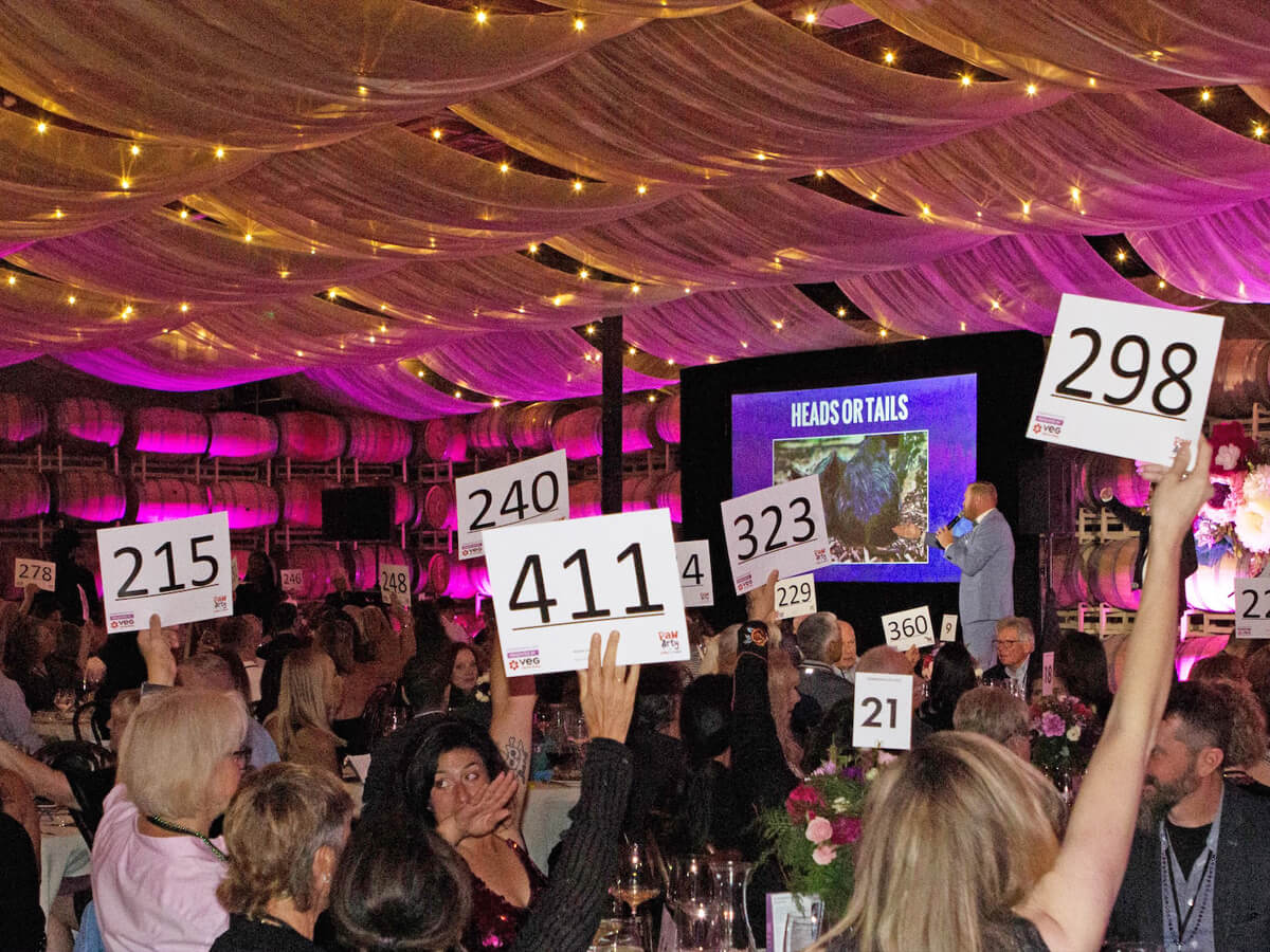 People seated at tables raise numbered paddles during Pasado's Celebration of Love event in a decorated venue with string lights and a stage.