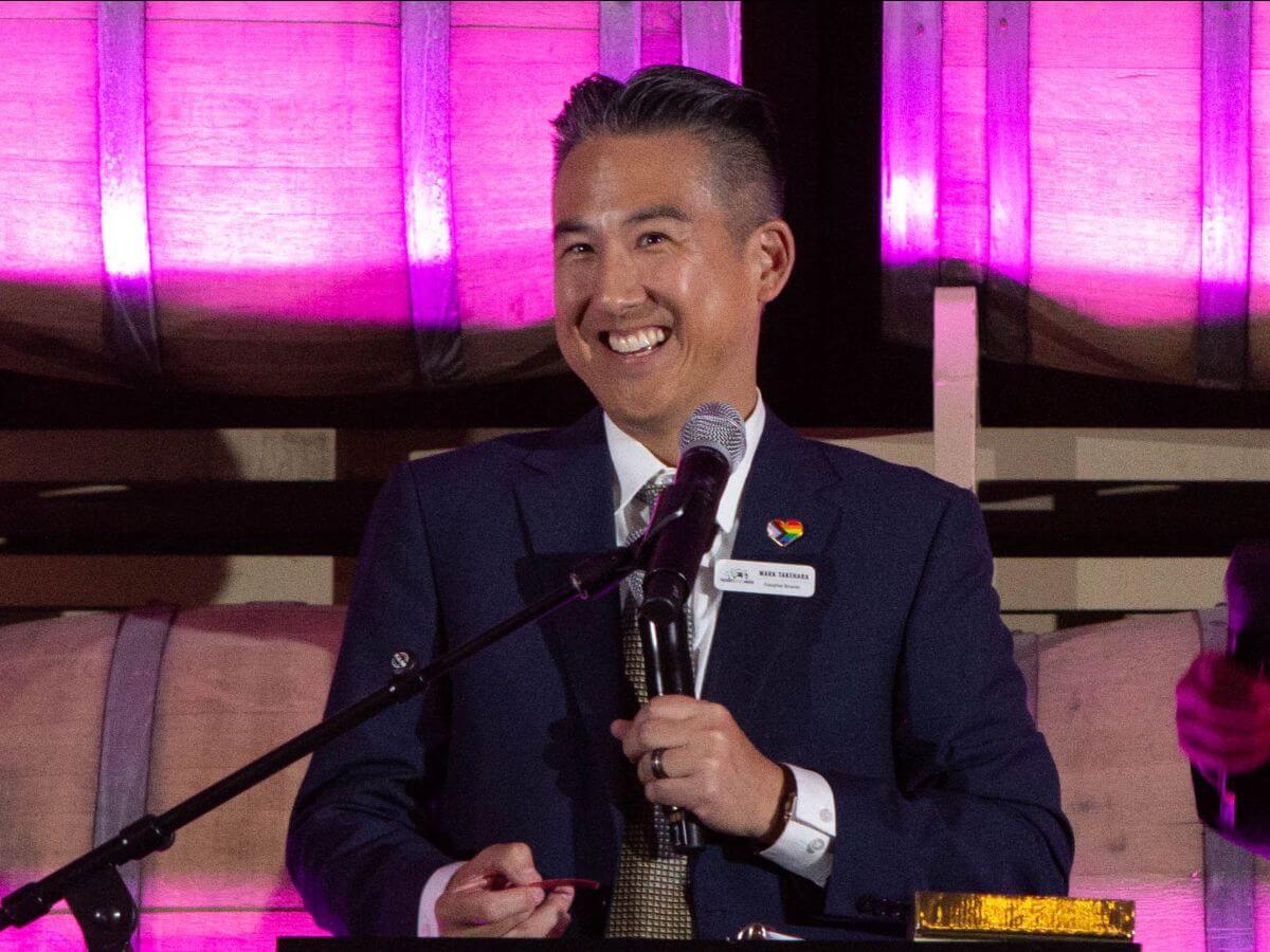 A person in a suit stands at a microphone, smiling, with wooden barrels and pink lighting in the background at Pasado's Celebration of Love event.