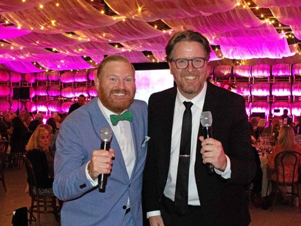 Two men in suits holding microphones smile at the camera during Pasado's Celebration of Love event, with a lively crowd and pink lighting in the background.