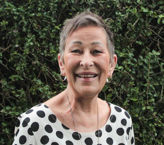 Awoman with short gray hair, wearing a white shirt with large black polka dots, smiles in front of leafy green bushes, ready to greet you as part of our friendly pet team.