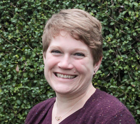 A person with short light brown hair, wearing a maroon sweater and a heart necklace, smiles in front of a leafy green hedge—ready to assist as part of the Pet Team.