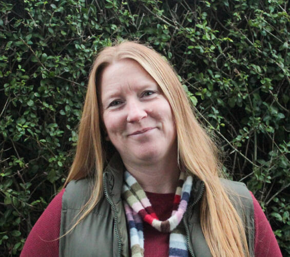 A woman with long red hair, wearing a multicolored scarf, red top, and green vest, stands in front of leafy green bushes and smiles at the camera—a proud member of the Pet Team.
