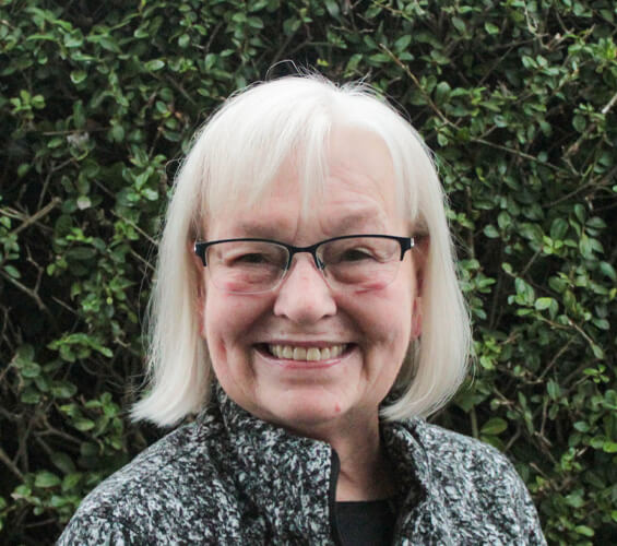 A woman with short white hair and glasses smiles in front of leafy green bushes, wearing a patterned jacket—ready to assist with friendly pet care as part of your local pet team.