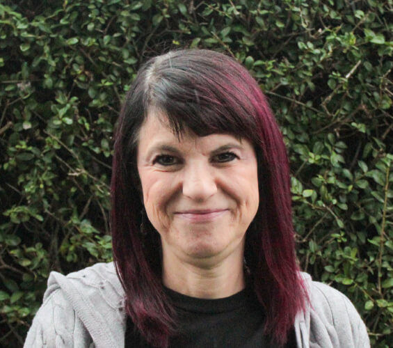 A member of the Pet Team with straight, shoulder-length dark hair with burgundy highlights stands outdoors in front of dense green foliage, wearing a grey hoodie and black top, smiling at the camera.
