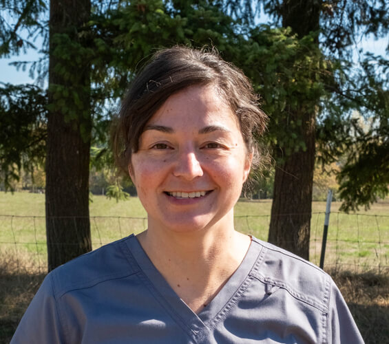 A person with short brown hair wearing gray scrubs stands outdoors in front of trees and a wire fence, smiling at the camera as part of the Pet Team.