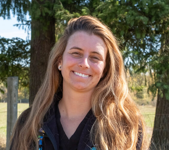A woman with long, light brown hair stands outdoors in front of trees, smiling at the camera. She is wearing a black top and jacket, ready to assist you as part of our dedicated Pet Team.