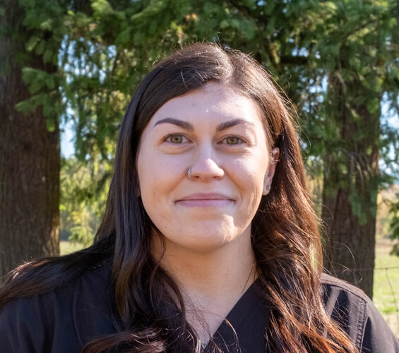 A woman with long dark hair and a black top stands outdoors in front of evergreen trees, looking at the camera—perfect for representing your trusted Pet Team in professional Pet Services.
