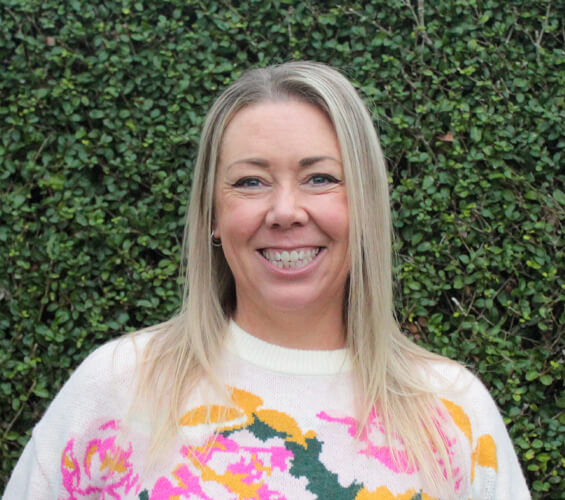 A woman with long blonde hair smiles in front of a green leafy hedge, wearing a white sweater with a colorful floral pattern—ready to provide friendly Pet Care as part of our dedicated Pet Team.