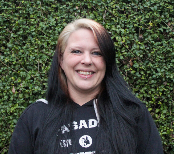 A woman with long black and blonde hair, wearing a black hoodie, stands smiling in front of a green leafy background, ready to represent top-notch Pet Care.