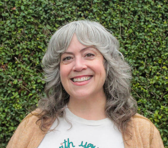 A smiling woman with gray, wavy hair stands in front of a green leafy background, wearing a white shirt and tan sweater—ready to provide expert pet care as part of our trusted pet team.