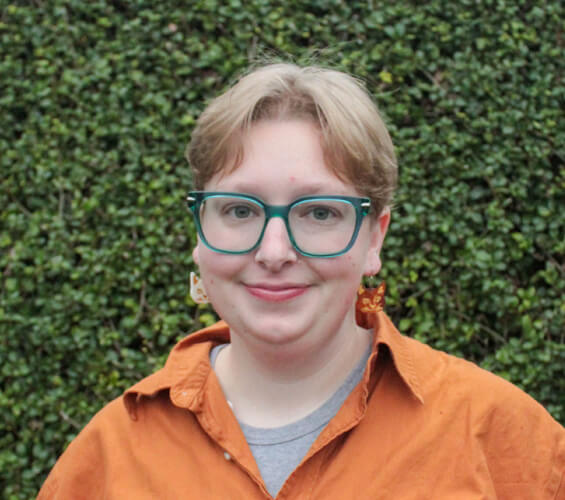 Person with short light hair, teal glasses, and an orange shirt stands in front of a leafy green hedge, wearing patterned earrings and smiling slightly—ready to bring warmth and care as a member of our dedicated pet care team.
