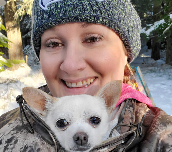 A woman in a knit hat smiles outdoors, holding a small white chihuahua partially zipped inside her camouflage jacket—showcasing the close bond fostered by dedicated Pet Services.